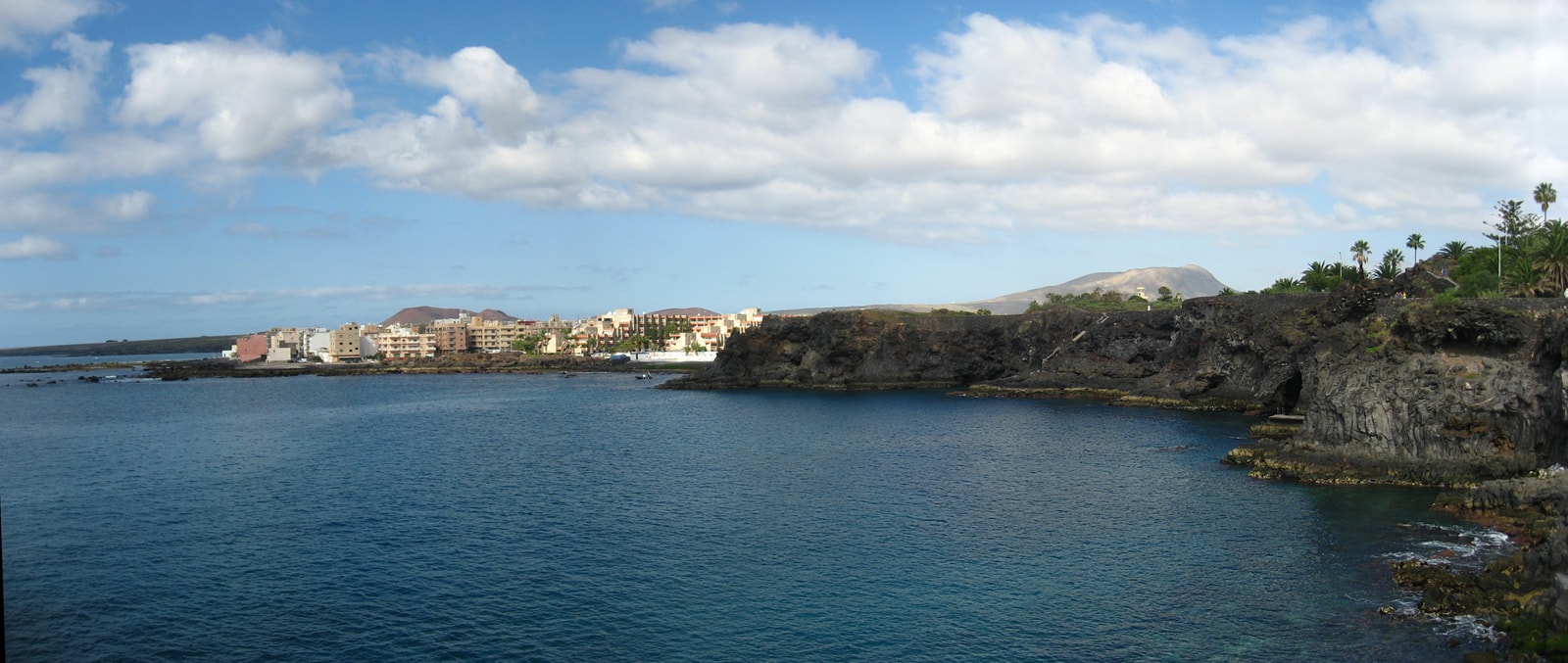 A sheltered bay with calm blue water is surrounded by dark volcanic coastline, with coastal buildings and a mountain in the background.