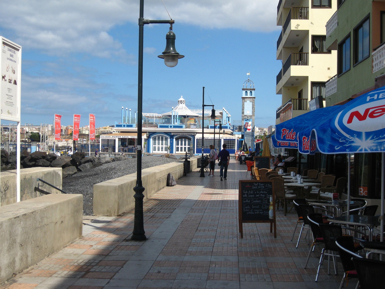A bustling harbor promenade with people sitting at outdoor cafes and walking along the waterfront on a sunny day.