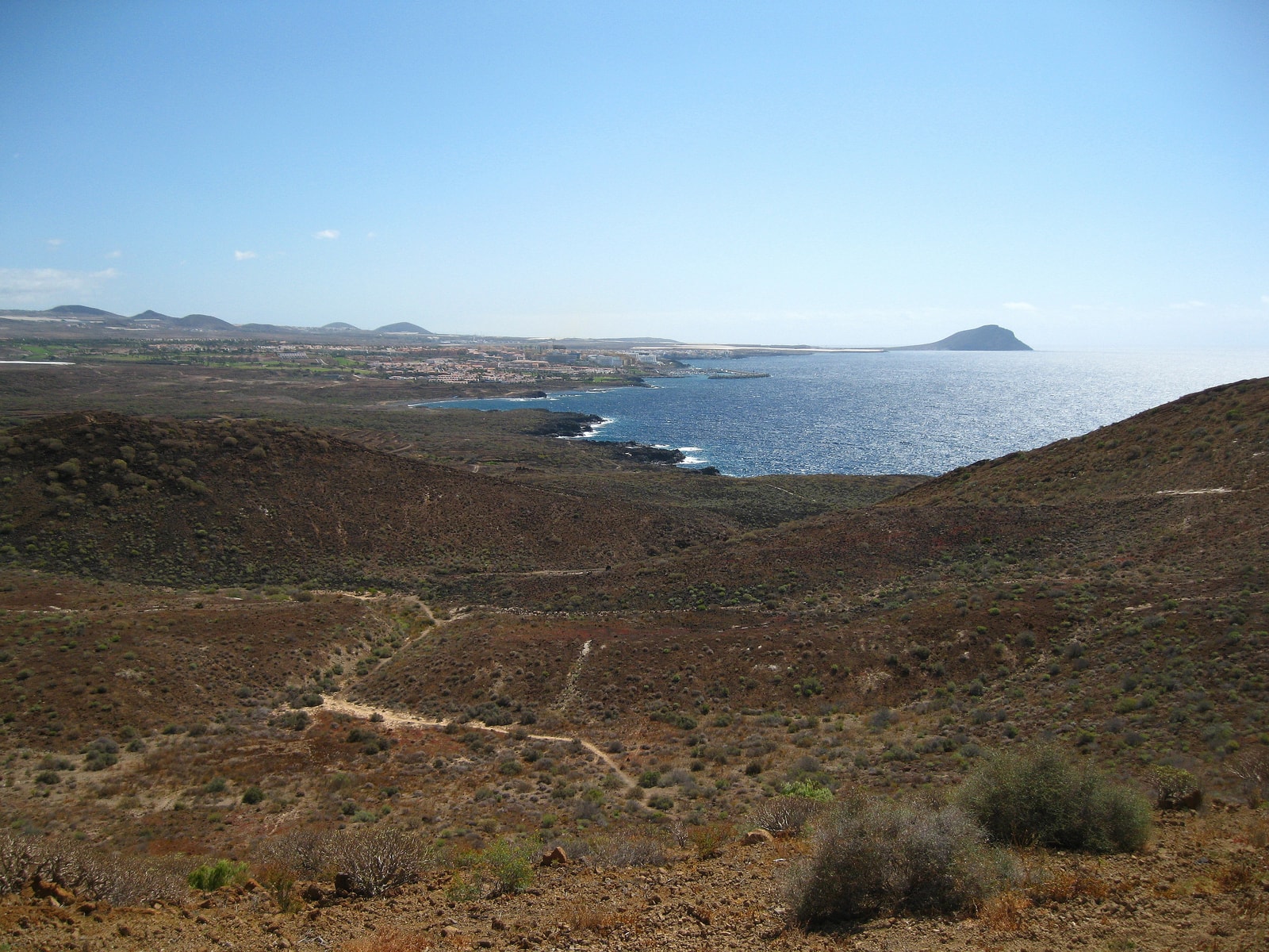 An expansive view of a rugged, arid coastal landscape with footpaths, volcanic hills, and the open sea.