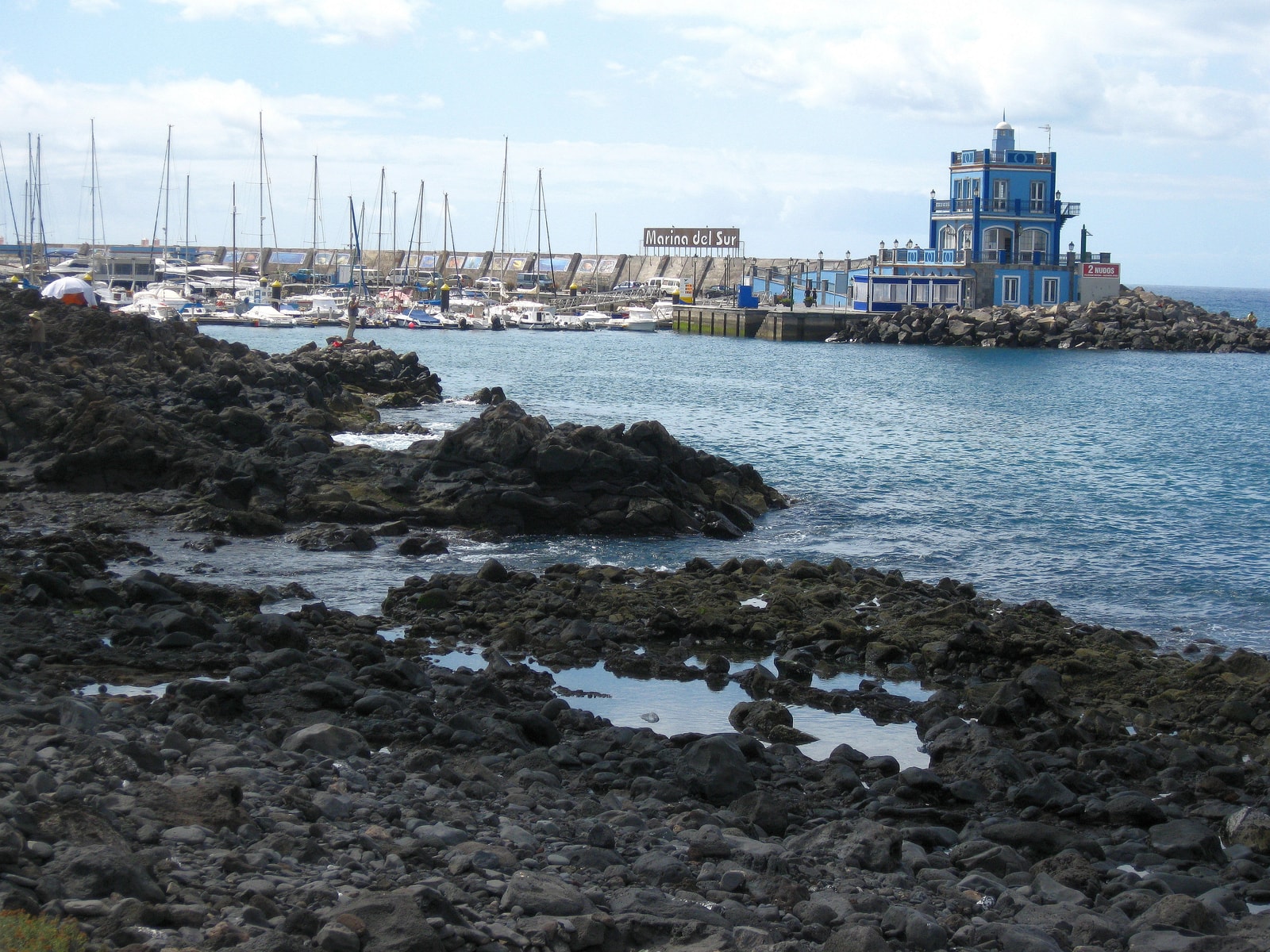 Dark volcanic rocks in the foreground with a marina full of boats and a breakwater building in the background.