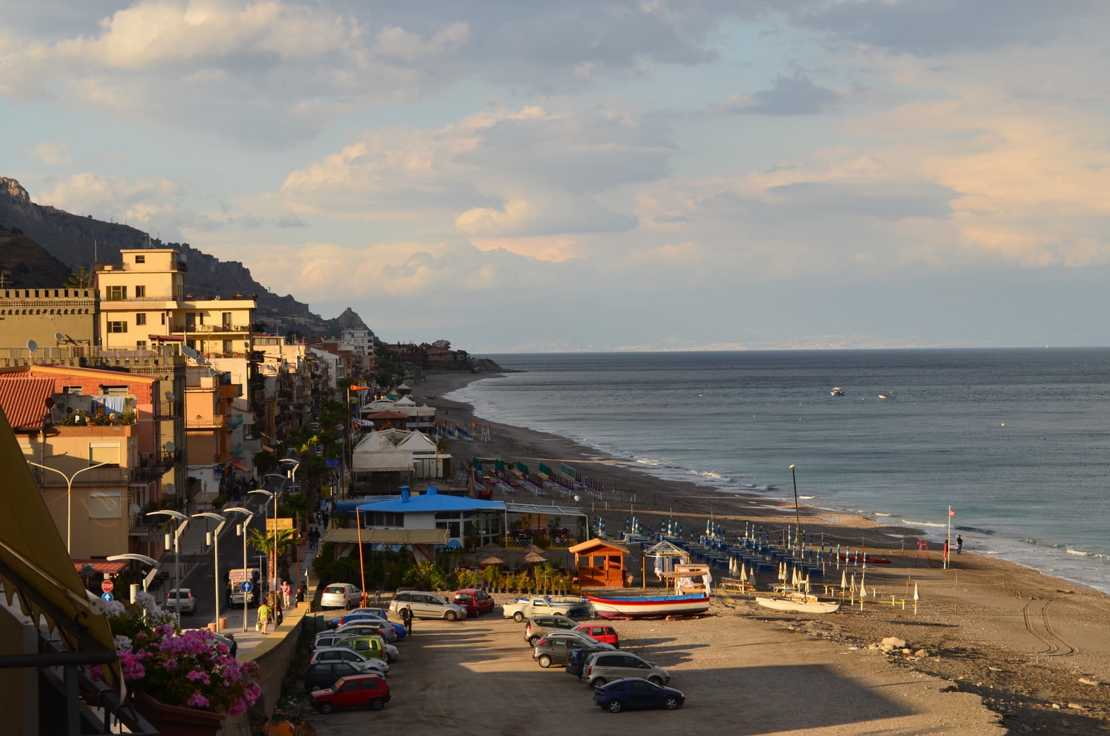The wide, sandy beach of Letojanni is pictured under a beautiful late afternoon sunset sky, with resort facilities visible along the shore.