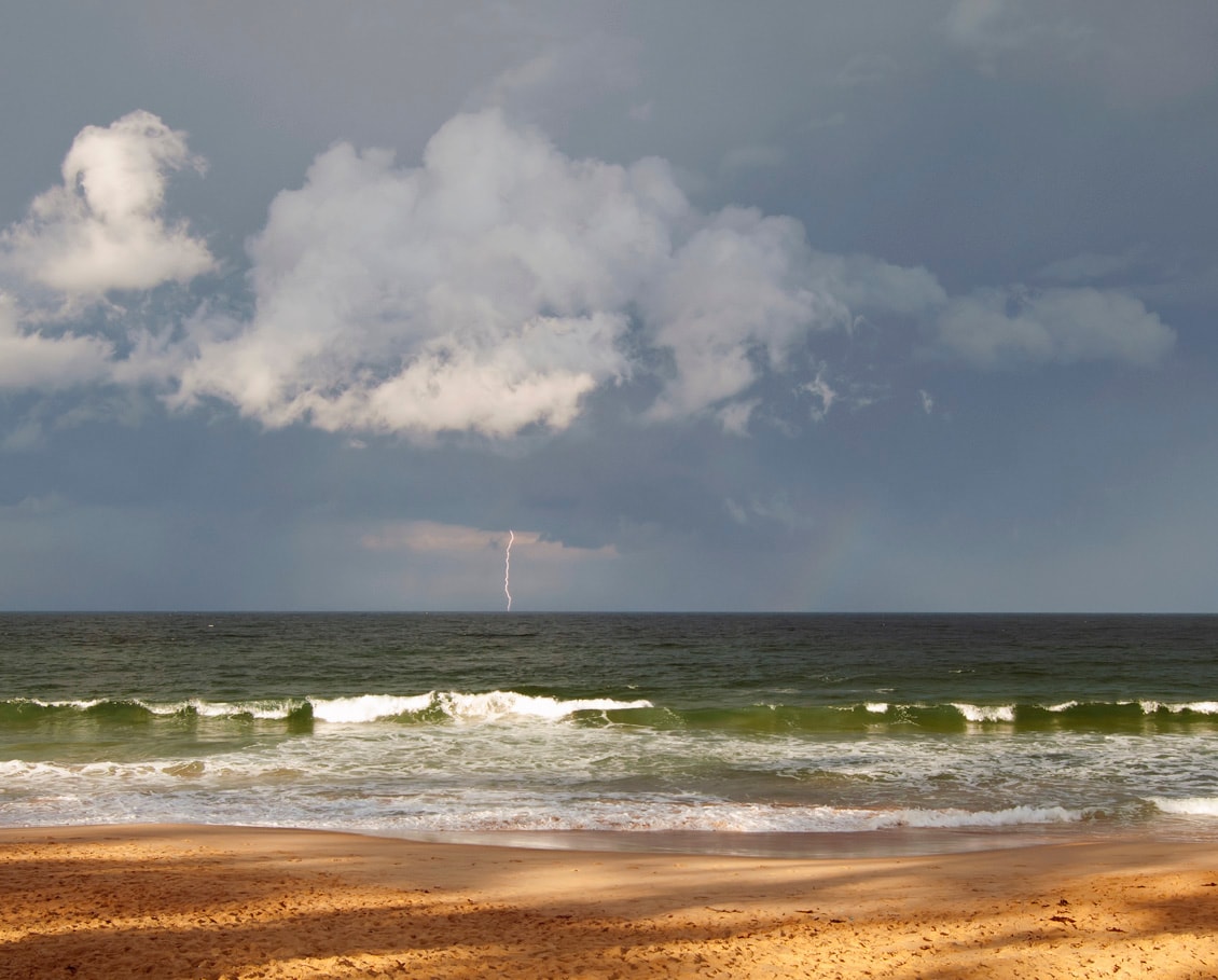 A dramatic bolt of lightning strikes over the ocean near Manly, illuminating the dark storm clouds and the coastline.
