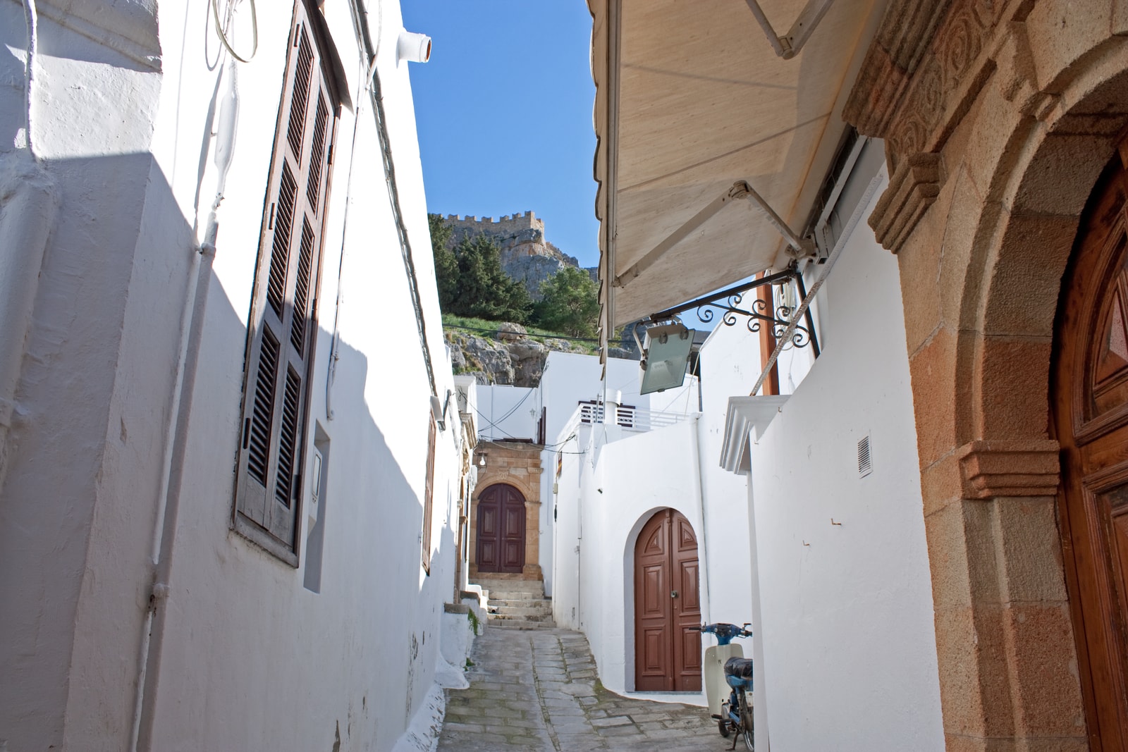 A narrow, whitewashed alley in Lindos village with stone paving leads towards the Acropolis in the background.