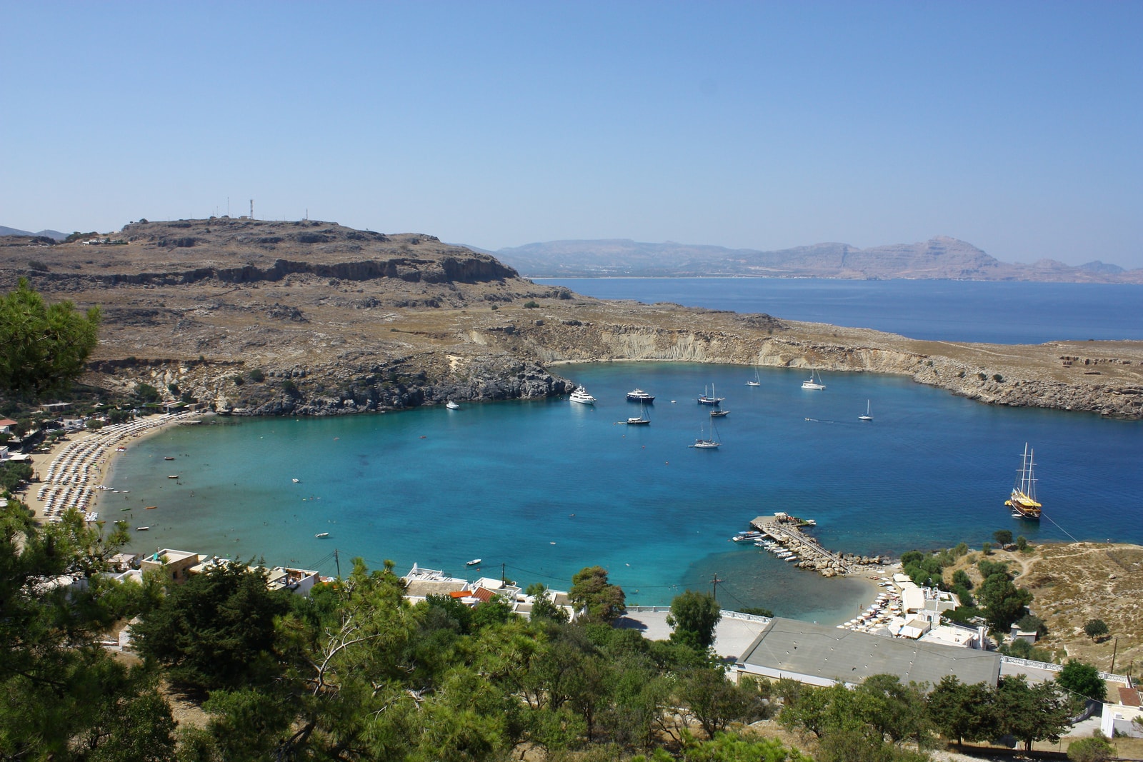 An aerial view of Lindos Bay, showing the sandy beach, turquoise water, and numerous boats anchored offshore.