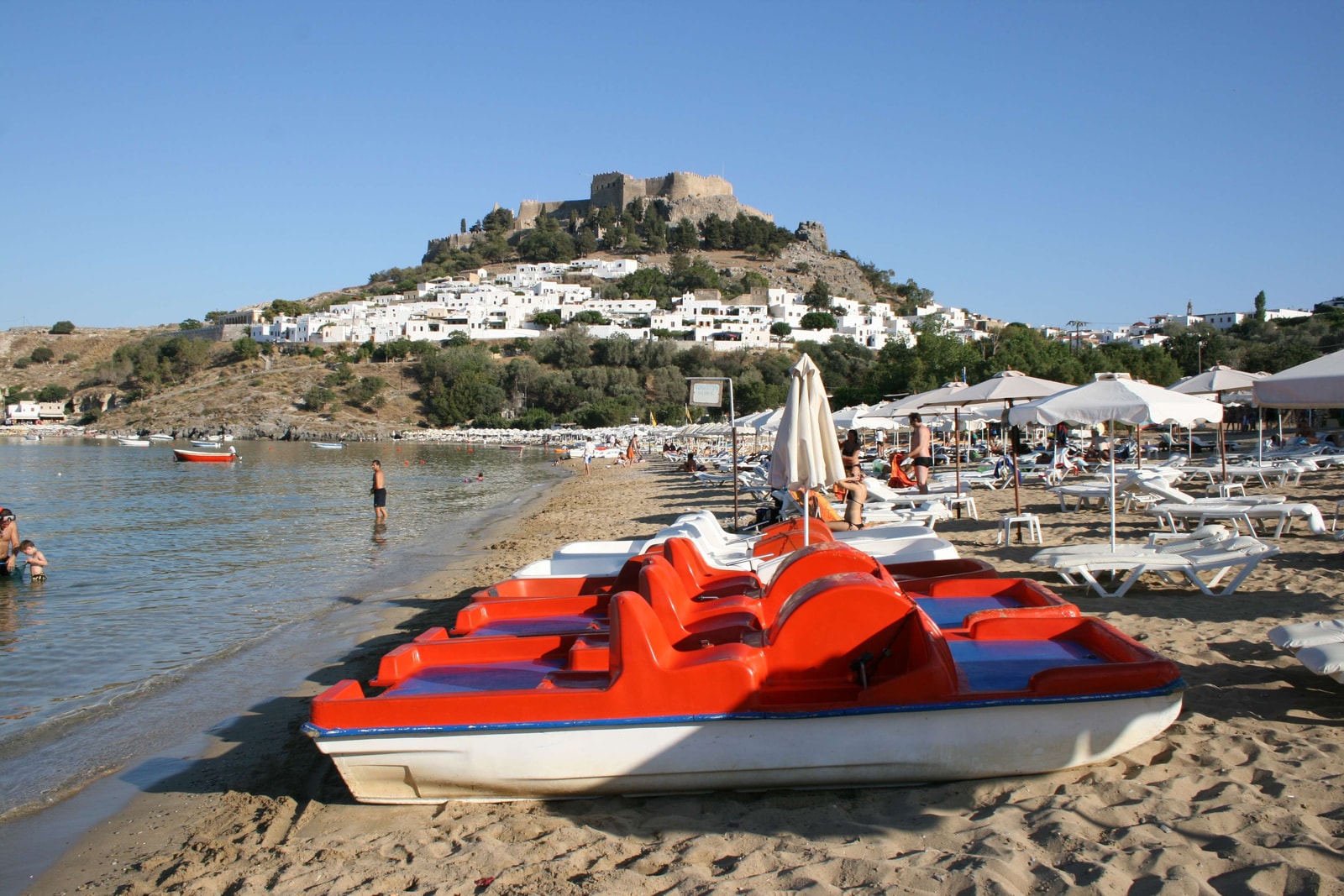 A bright red pedal boat sits on the sand at Lindos Beach, with the Acropolis visible on the hill behind it.