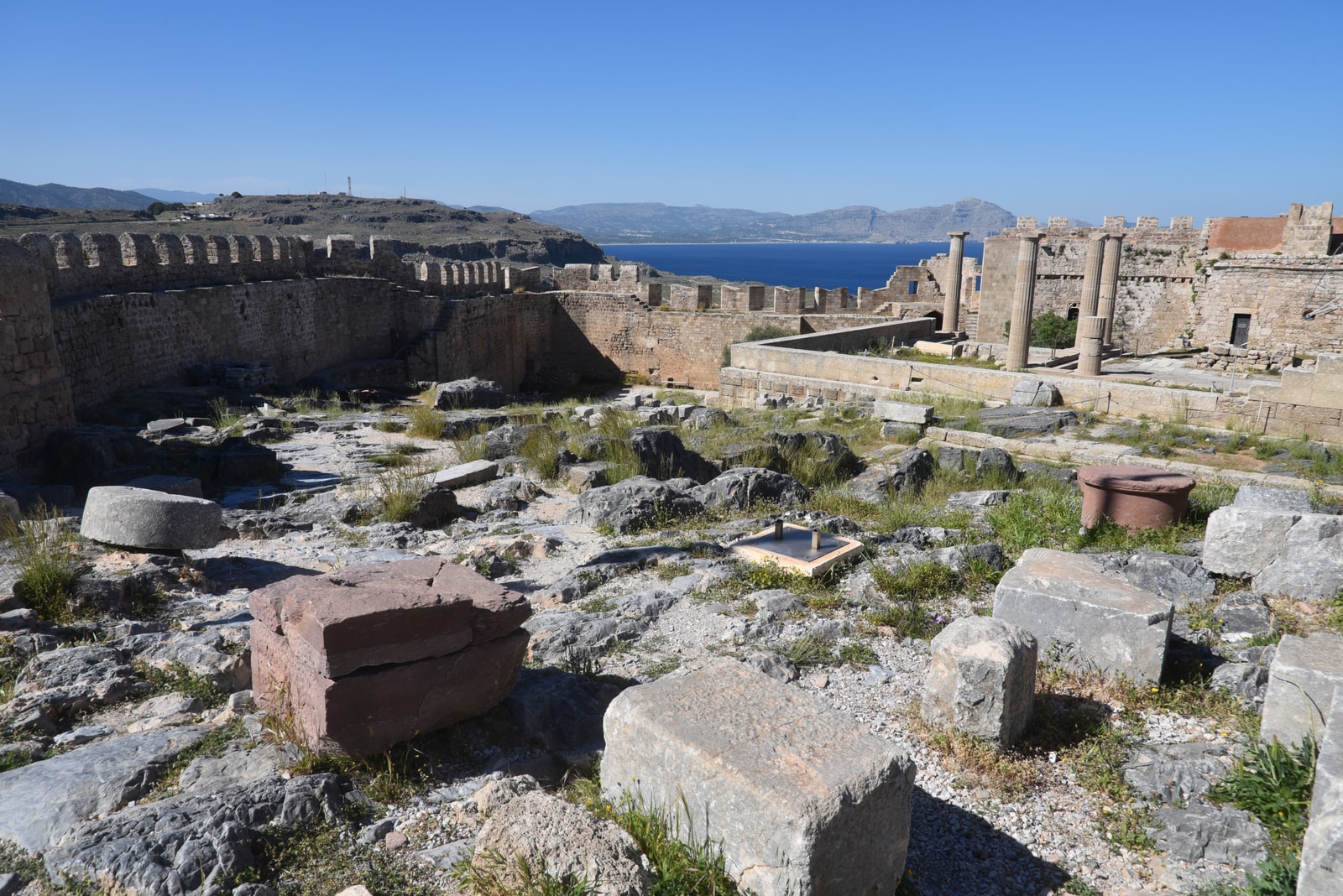 A view of the Acropolis of Lindos perched on a cliff above a turquoise bay with a small beach and several boats.