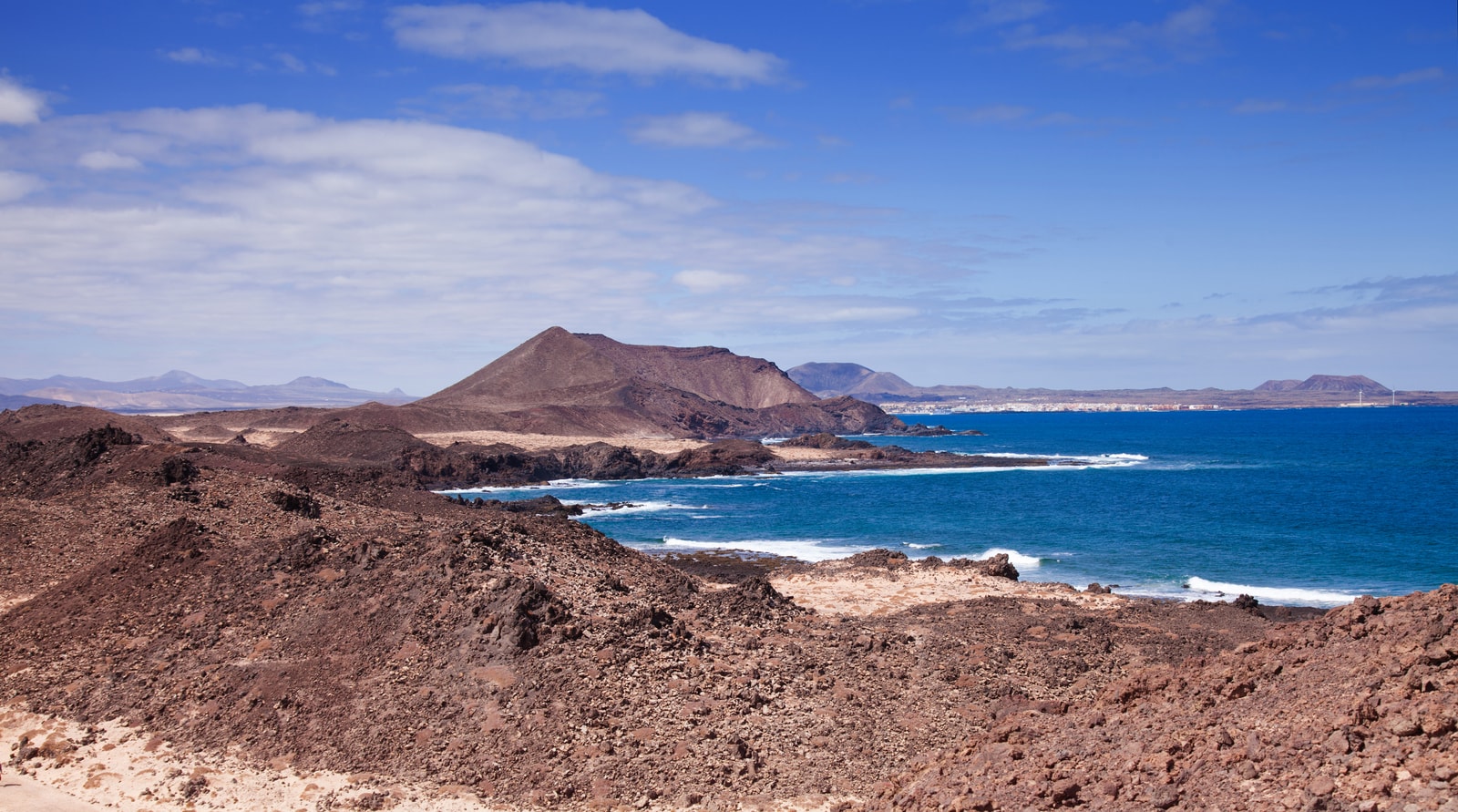 A rugged volcanic coastline on Lobos Island with the town of Corralejo visible across the water in the background.