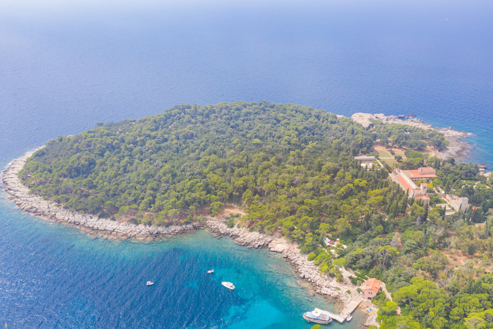 An aerial view of a wooded island with a historic monastery and a small harbor with boats.