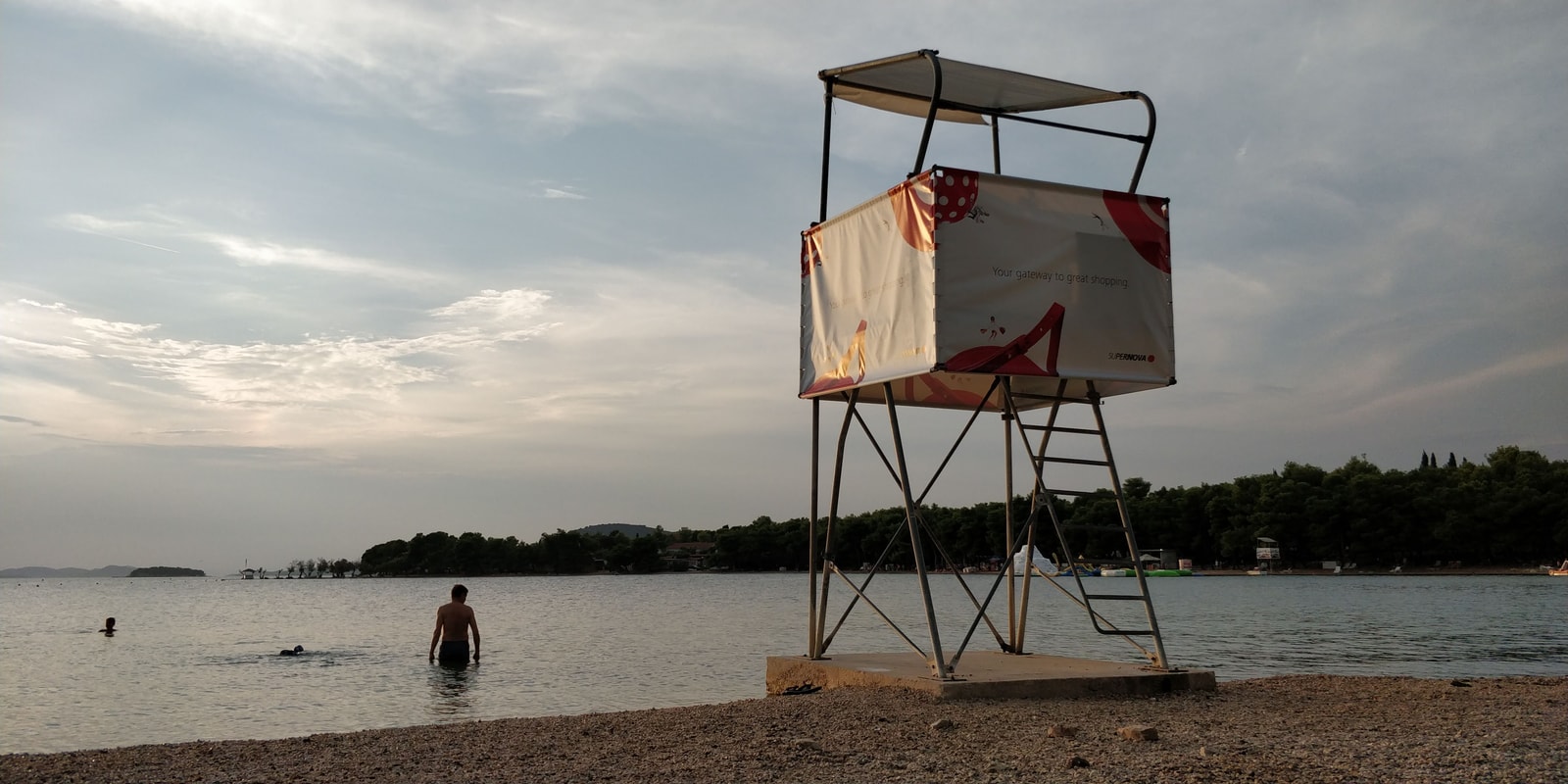 The pebble shoreline and clear, calm water of Lolić Beach in Pirovac, with a lifeguard tower visible.