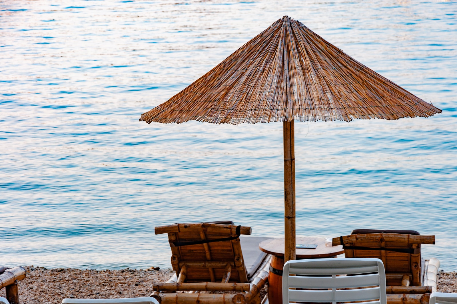 Natural-textured bamboo lounge chairs and a thatched straw umbrella sit on the smooth white pebble shore of Lone Beach.
