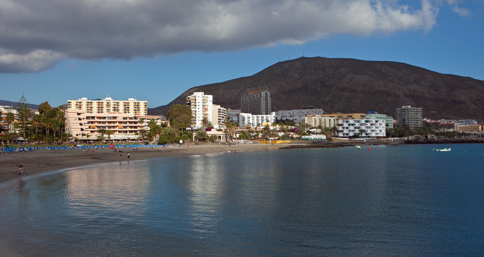 A wide sandy beach curves around a calm blue bay, with a resort town built into the hillside and a dark volcanic mountain in the background.
