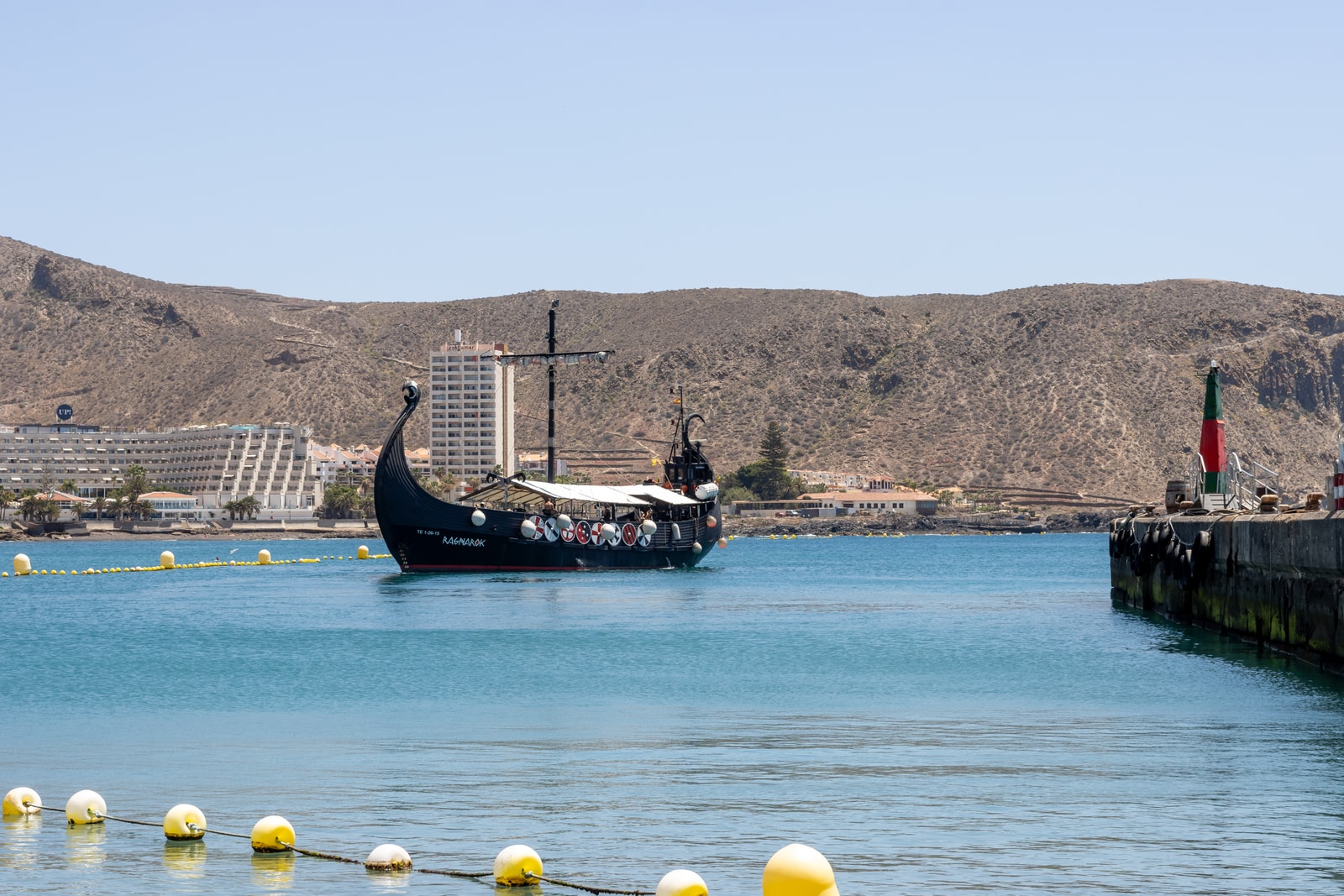 A distinctive Viking-style excursion boat sails through a turquoise harbor, with a dry volcanic hillside in the background.