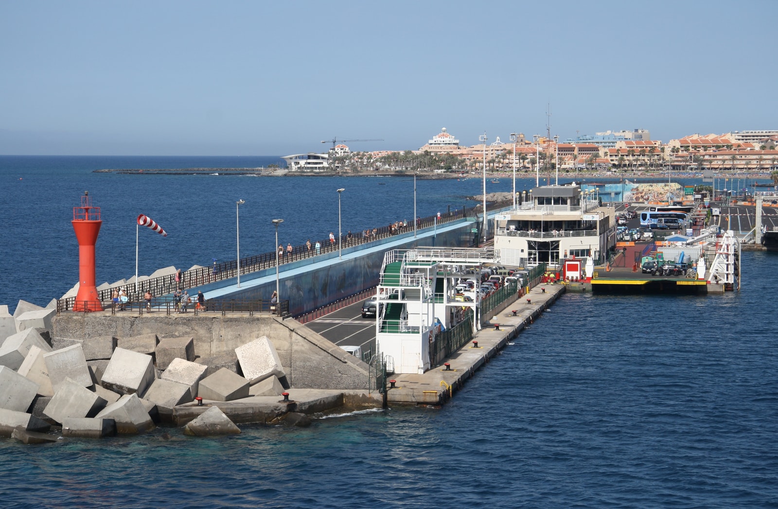 A view from a ferry showing a long breakwater with a red lighthouse at the end, protecting the harbor of Los Cristianos.