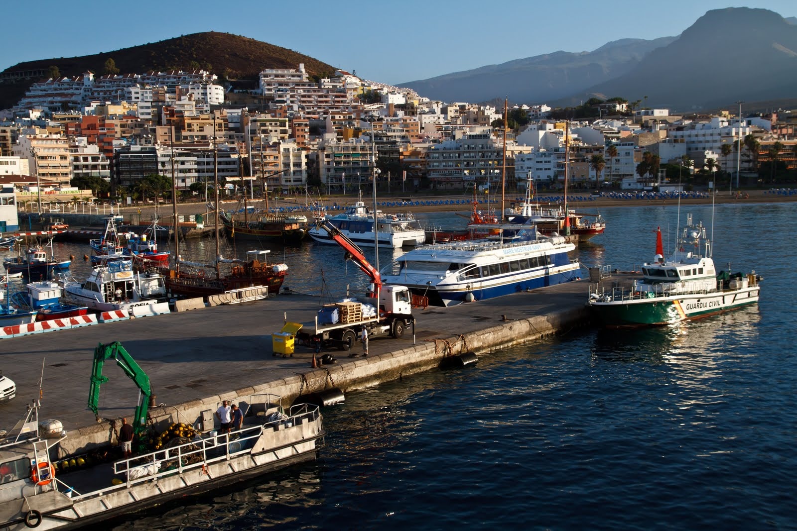 A harbor scene with fishing boats, a pier, and the town of Los Cristianos built on the hillside under warm light.