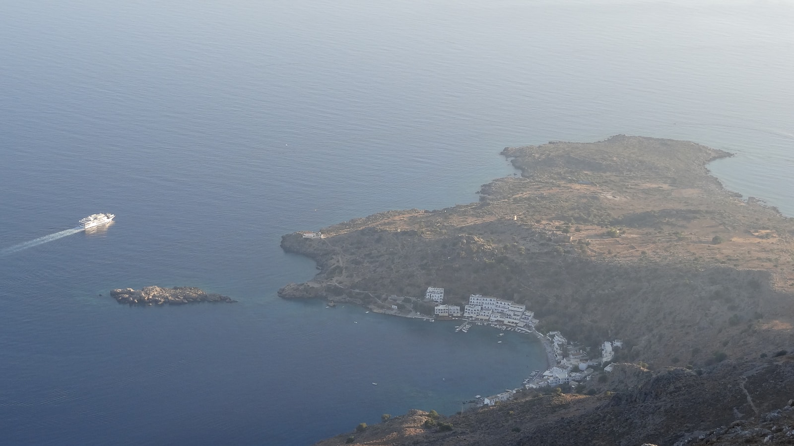 A high-angle view shows the white-washed village of Loutro wrapping around a perfectly calm, emerald-green bay.
