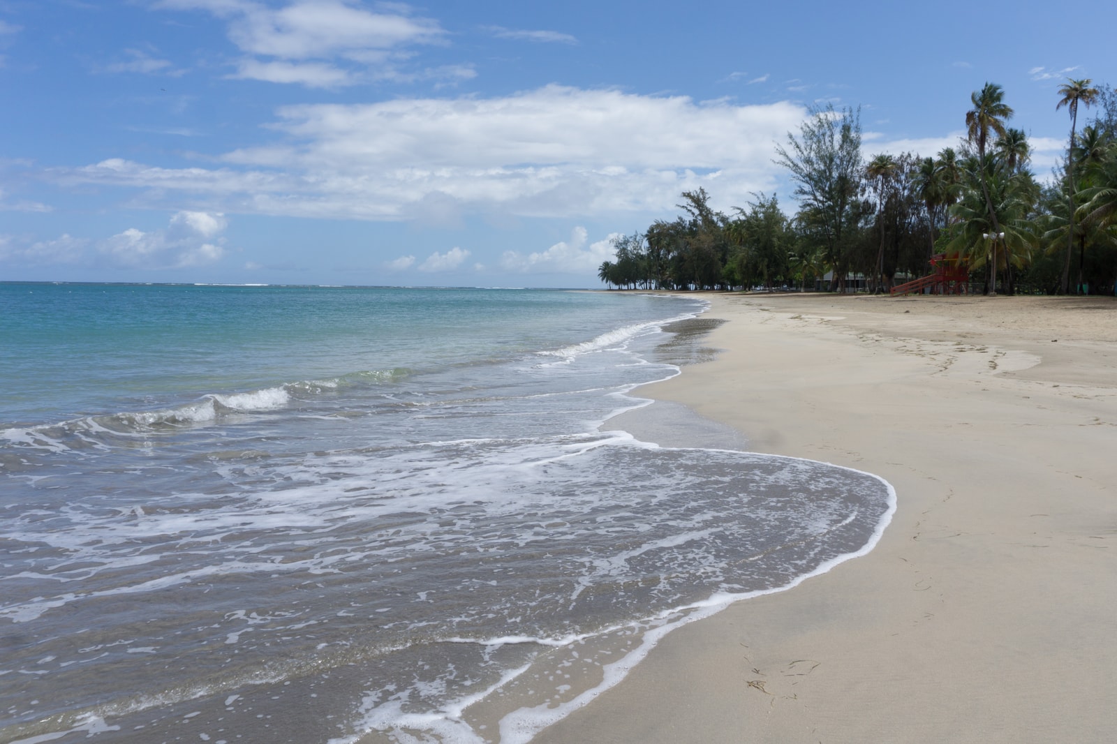 A wide, curving shoreline with light sand, turquoise water, and a line of palm trees in the distance.