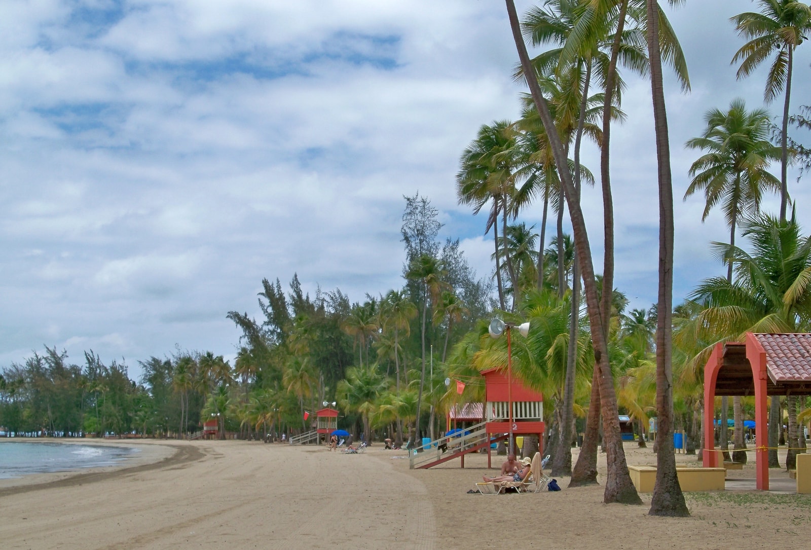 A view of Luquillo Beach showing palm trees, a lifeguard station, and covered picnic shelters near the sand.