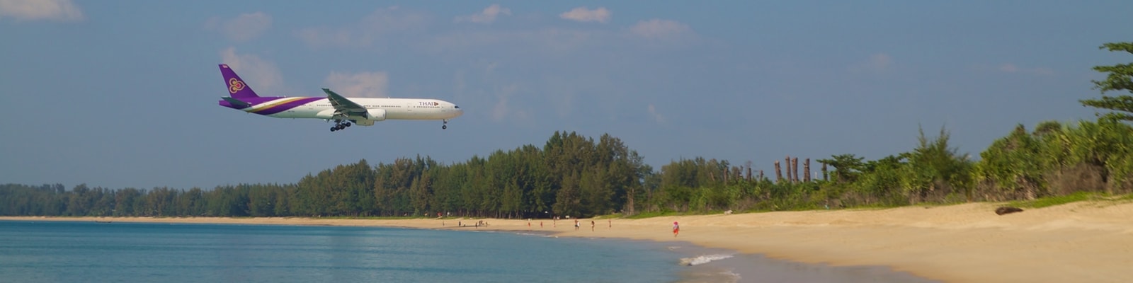 A large passenger airplane flies low over the water as it approaches the runway near Mai Khao Beach.