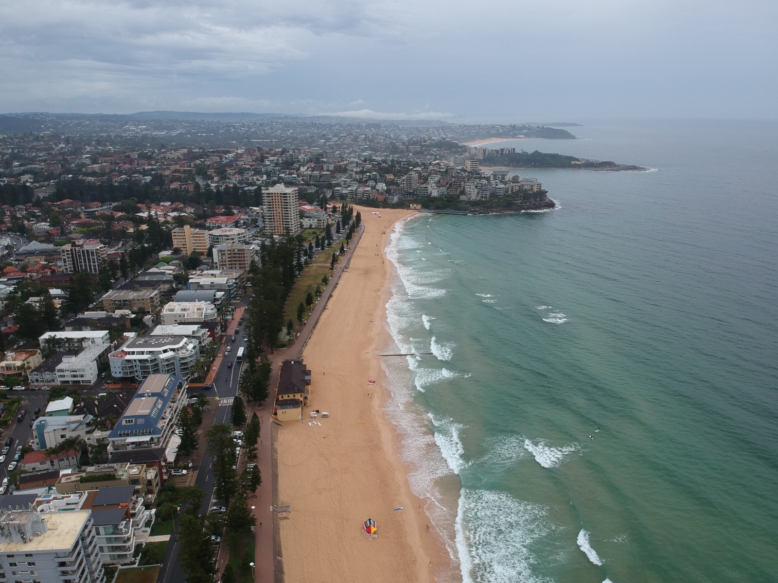 An aerial view of Manly Beach shows a long, wide sandy shore with lines of white surf rolling in, bordered by a dense town and green parkland.
