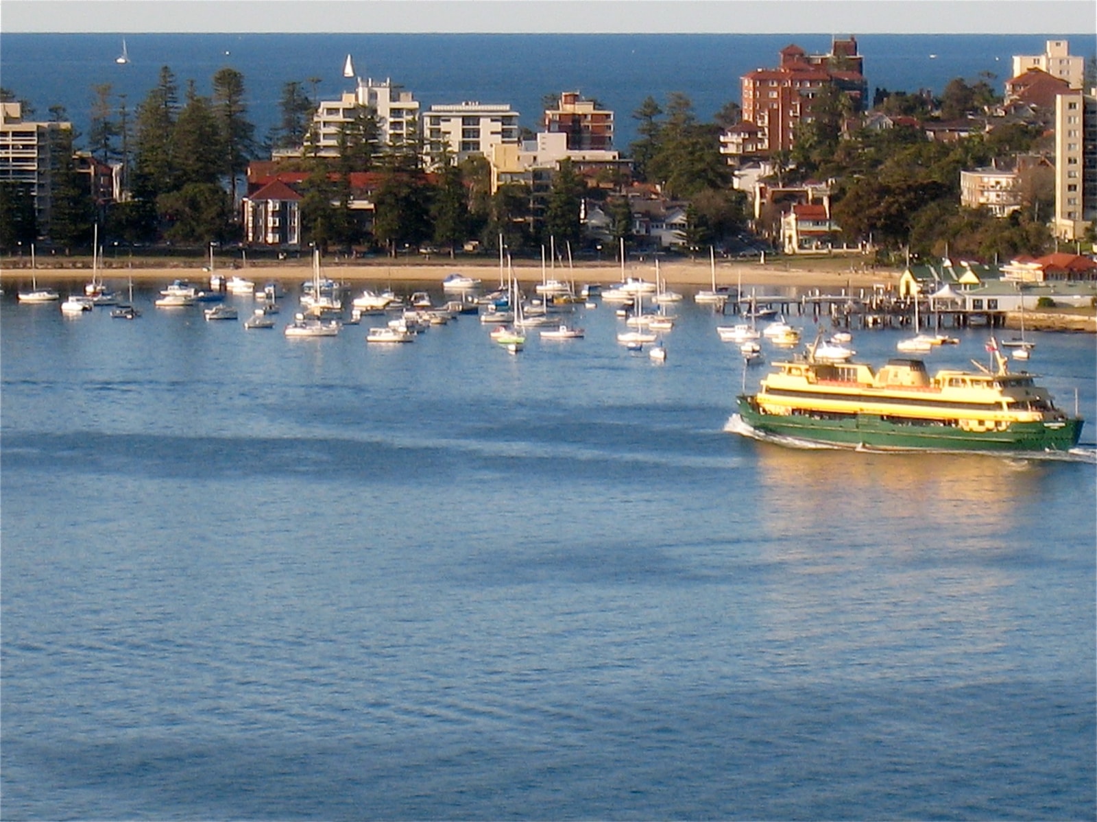 A Sydney ferry approaches the wharf at Manly, with sailboats moored in the calm harbour waters in the foreground.