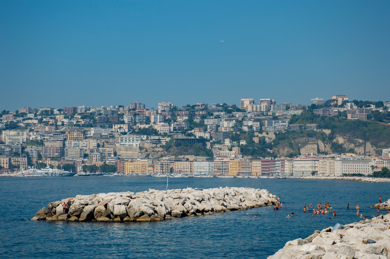 Swimmers enjoying the water at Mappatella Beach along the Lungomare Caracciolo, with a rocky breakwater in the foreground and colorful coastal buildings behind.