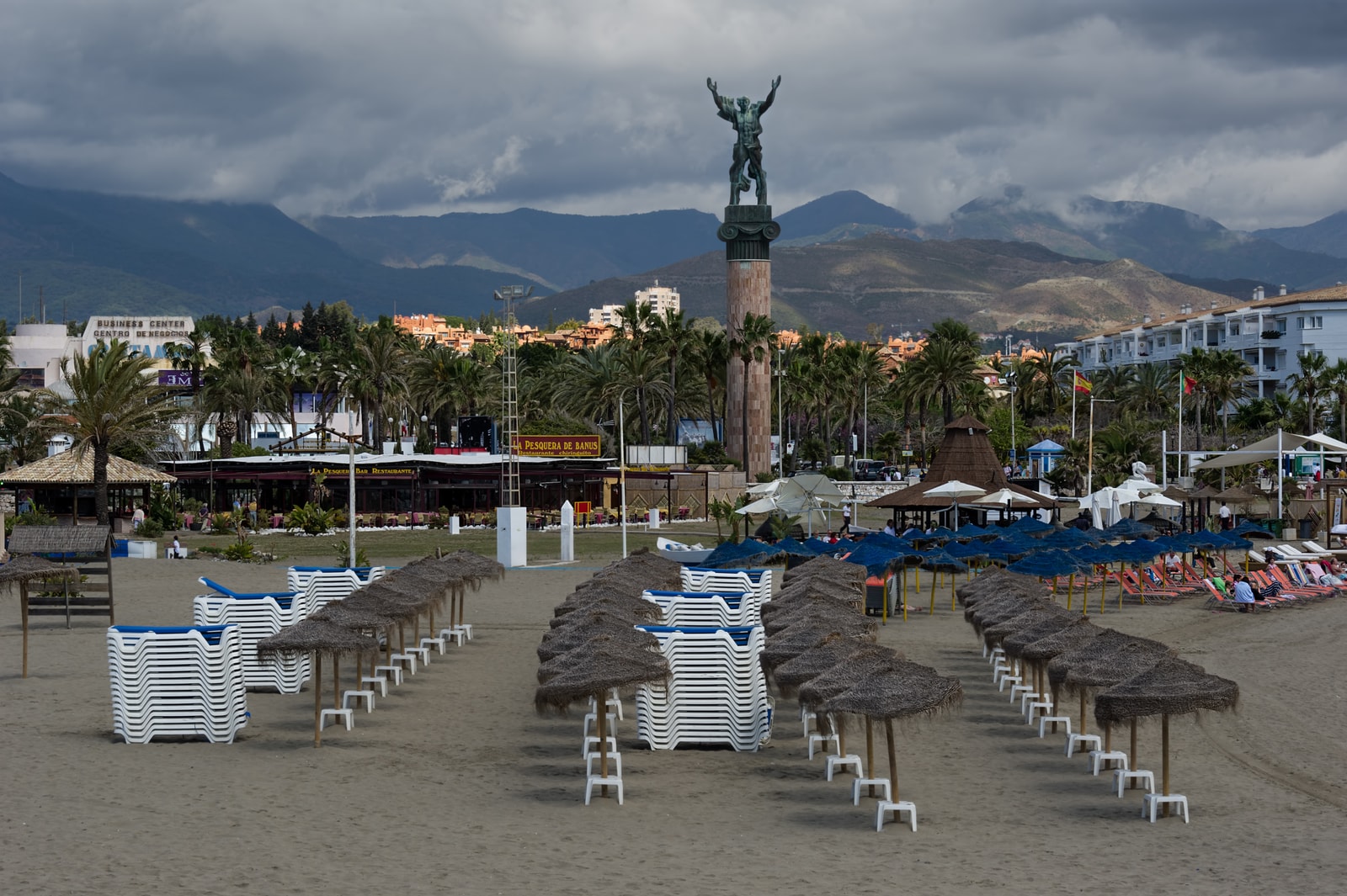 A view of stacked lounge chairs on a Marbella beach during the off-season, with the iconic column statue near Puerto Banús against a misty mountain backdrop.