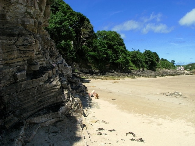 A sunny day at a sandy cove, with calm blue water, green hills in the background, and people enjoying the beach.