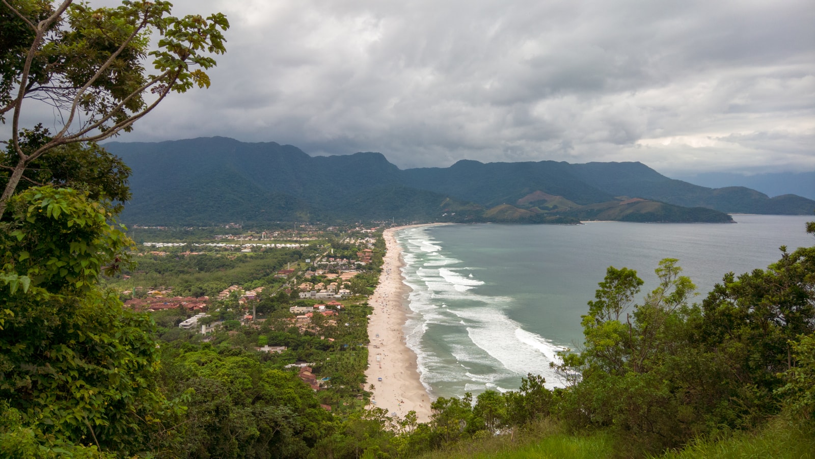 A sweeping aerial view of Maresias beach, showing its long sandy shore, the surrounding green mountains of the Atlantic Forest, and the town nestled along the coast.