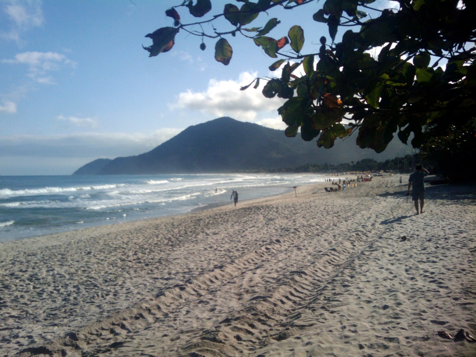 A ground-level view of Maresias beach, showing waves, beachgoers, and the prominent mountain backdrop covered in green vegetation.