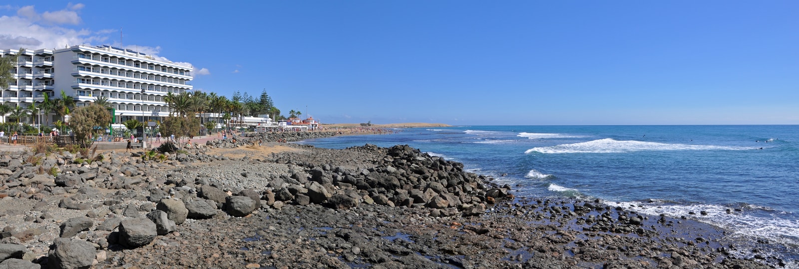 A panoramic view of the Maspalomas coastline shows turquoise water, a sandy beach, and luxury resort buildings along the shore.
