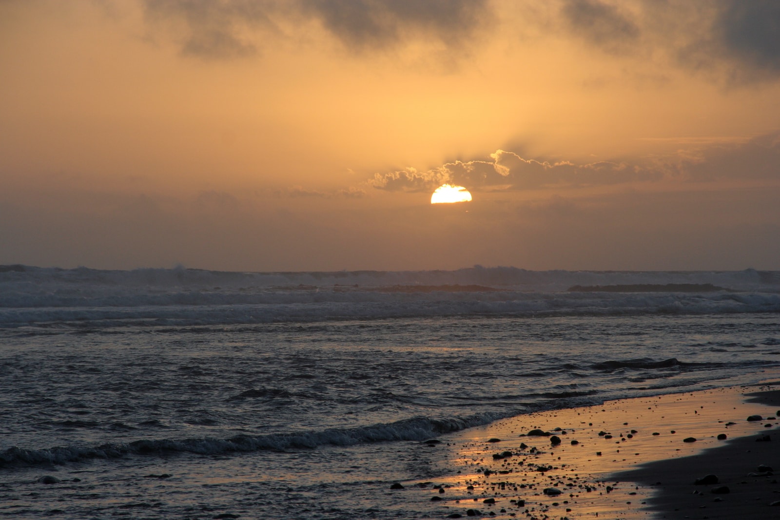 The sun sets over the ocean, casting a golden glow on the wet sand of the beach.