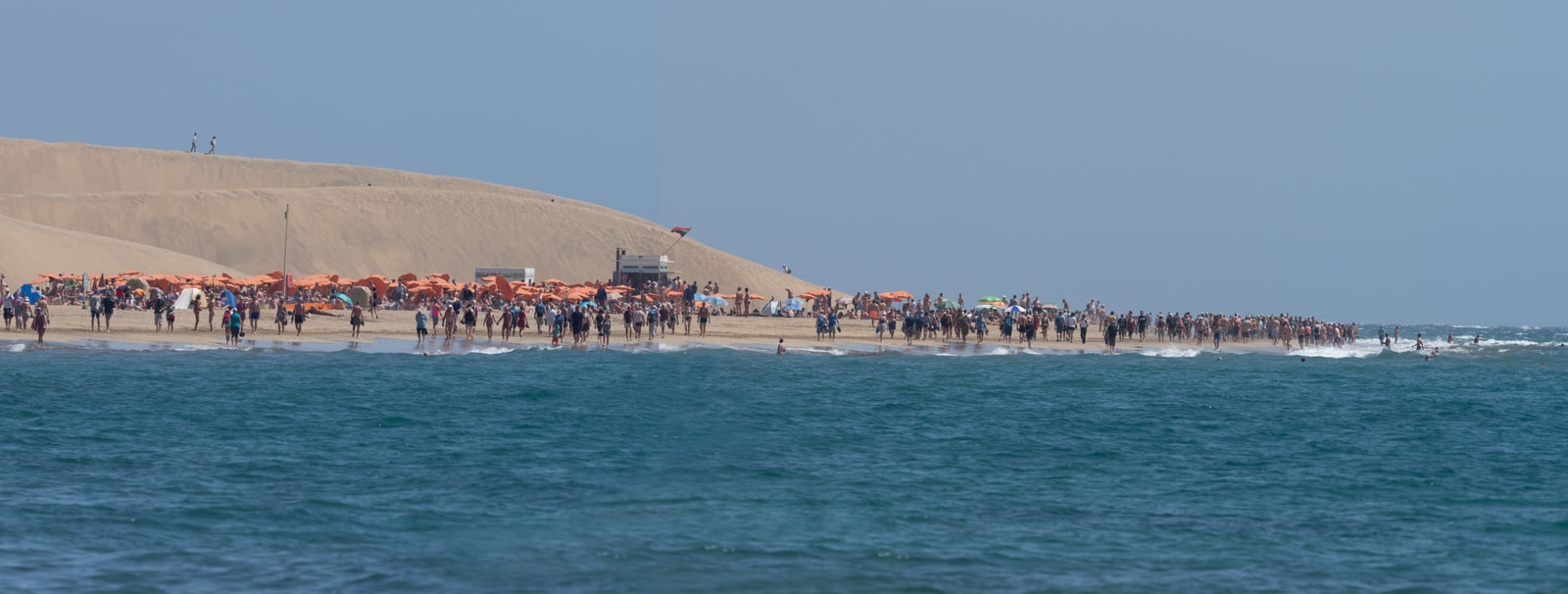 A wide panoramic shot of Maspalomas beach, filled with colorful parasols and beachgoers enjoying the sun, with the vast dunes in the background.