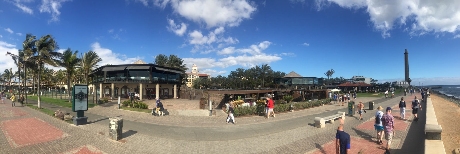 A panoramic photo shows a bustling pedestrian walkway lined with upscale shops and restaurants, with the lighthouse visible in the distance.