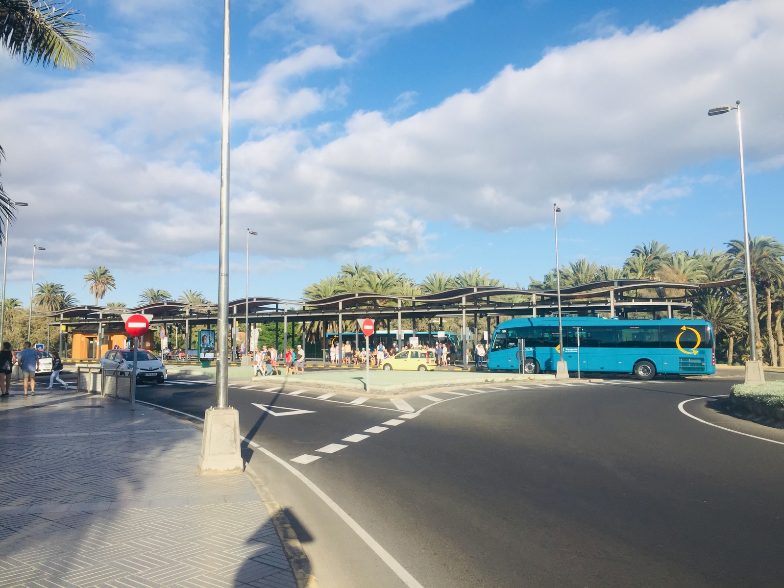 A modern, open-air bus station with blue public buses parked under a canopy, surrounded by palm trees.