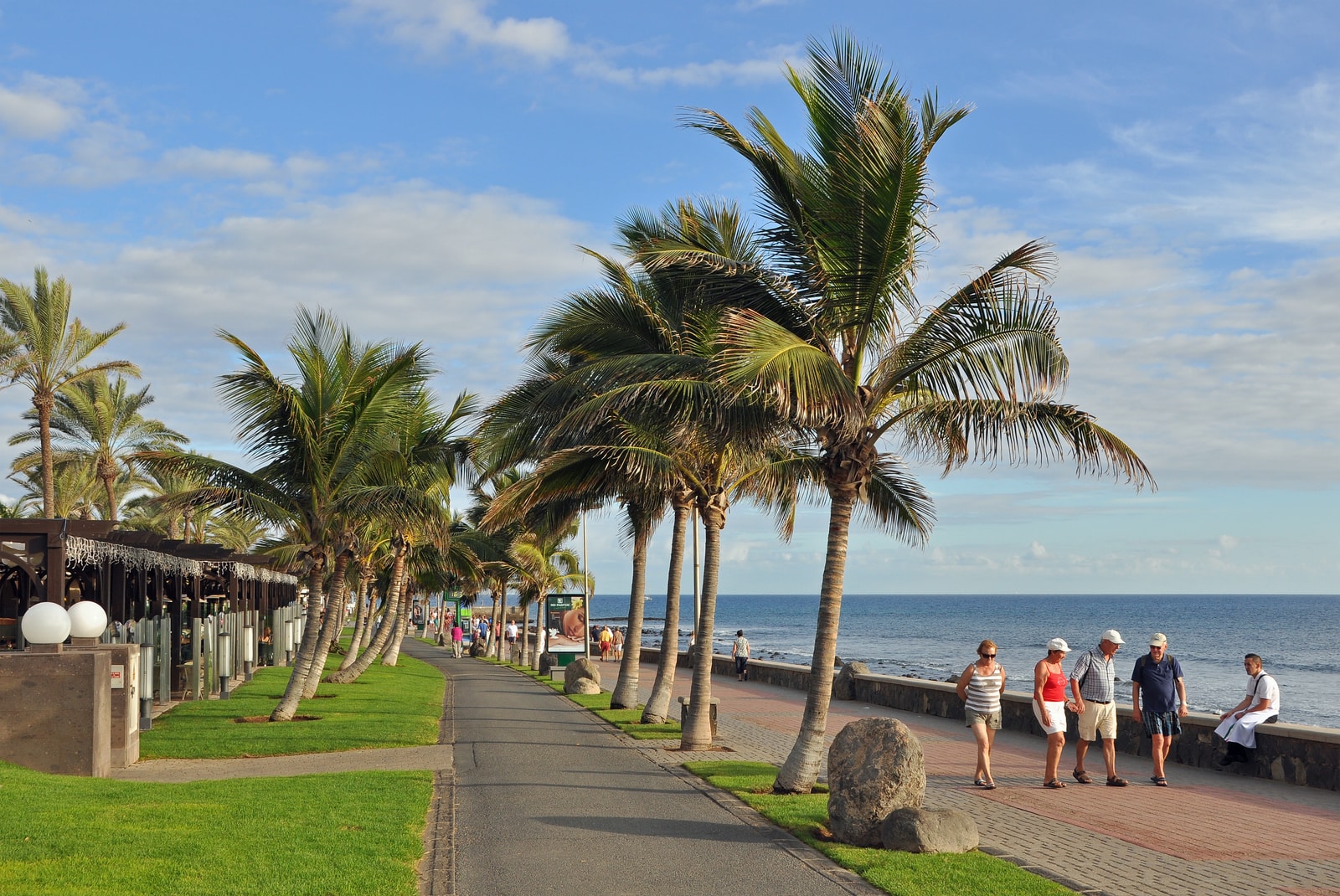 A wide, brick-paved promenade lined with tall palm trees runs alongside the ocean, with elegant resort buildings in the background.