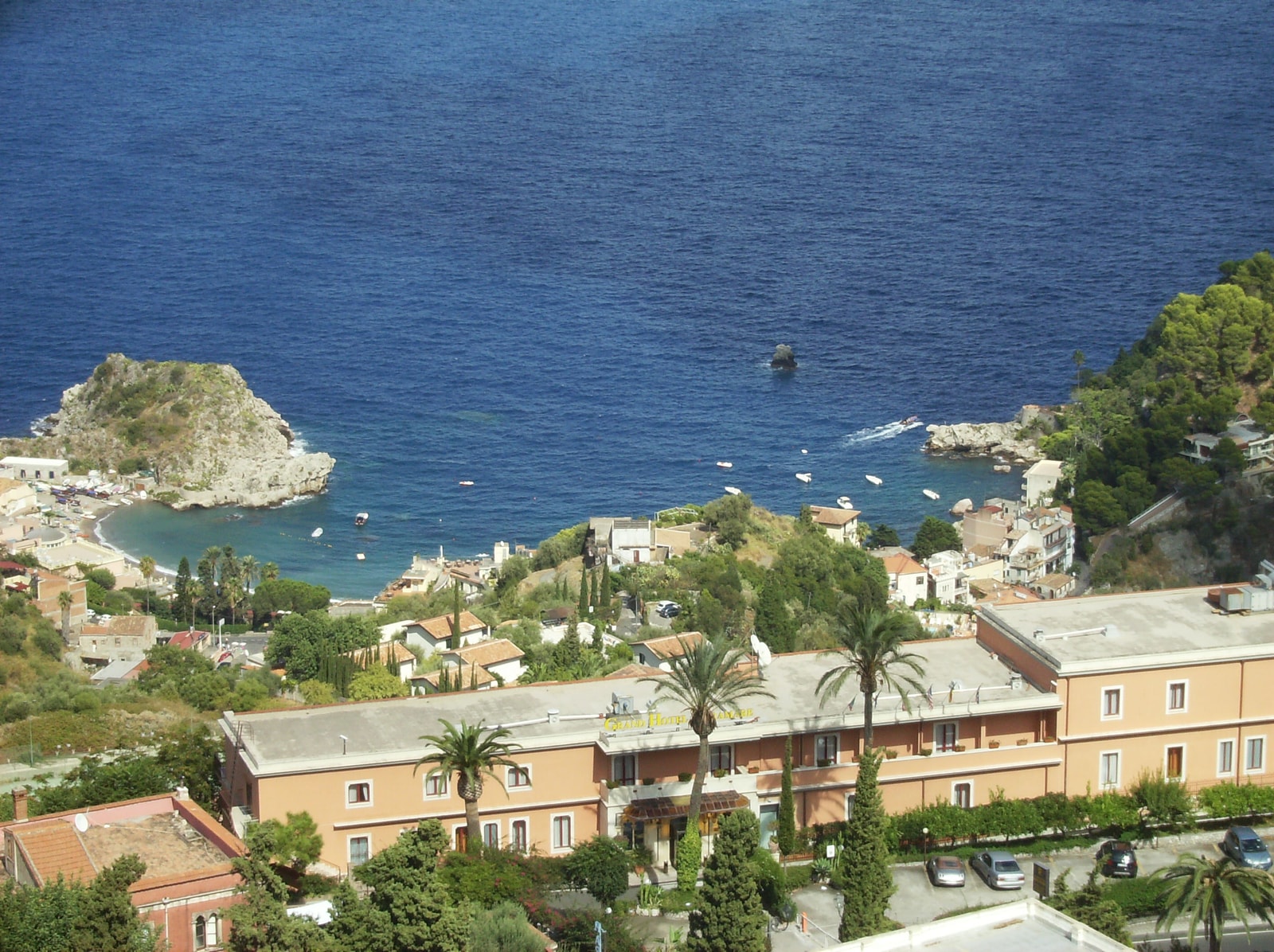 A high-angle overview of the cove of Mazzarò, showing its beach, coastal architecture, and the boats that offer tours of the nearby sea caves.