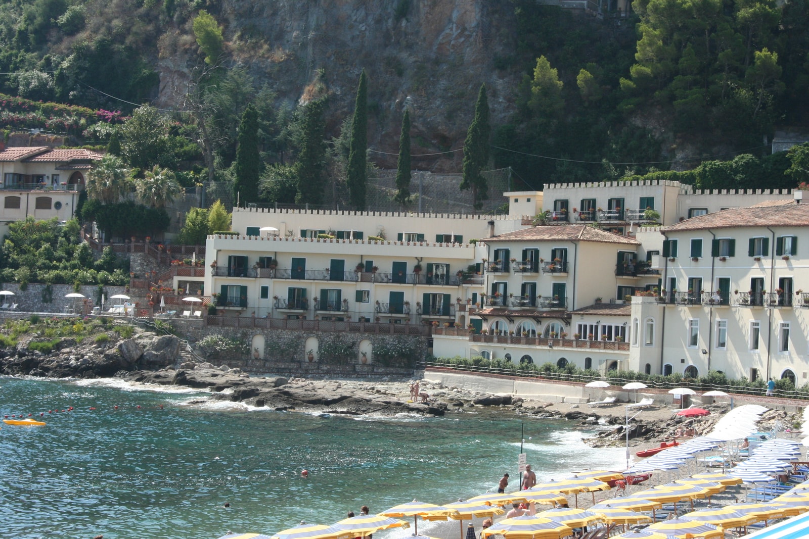 A stylish view of Mazzarò Beach, featuring colorful umbrellas and an elegant hotel designed with battlements, reflecting the area's luxury resort atmosphere.