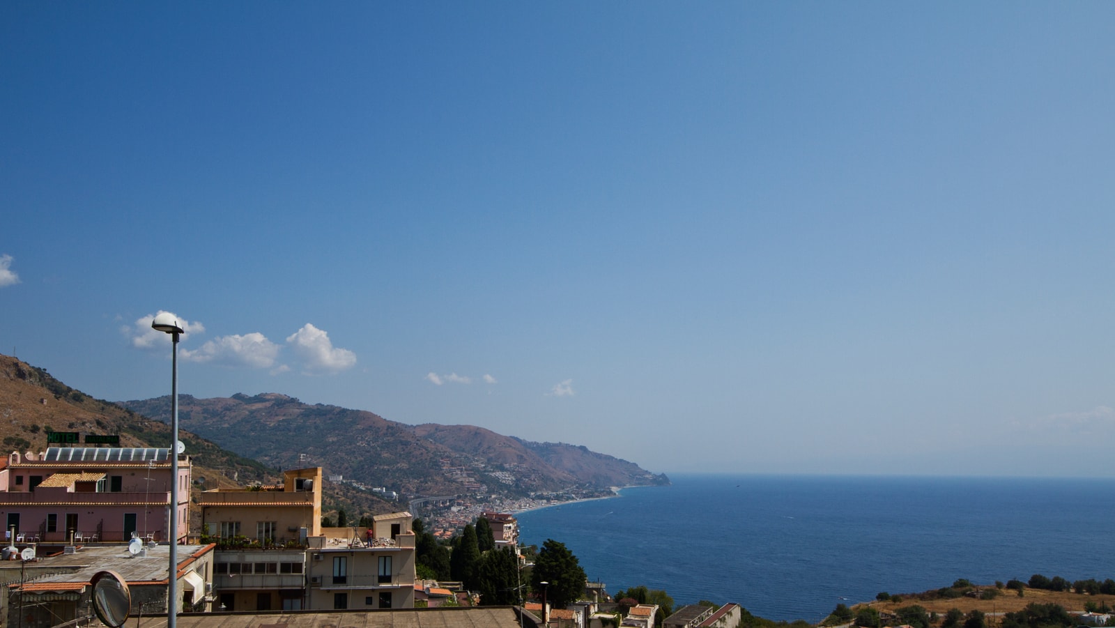An elevated, panoramic view of Mazzeo beach shows its long, sandy shoreline stretching along the coast toward the hills in the distance.
