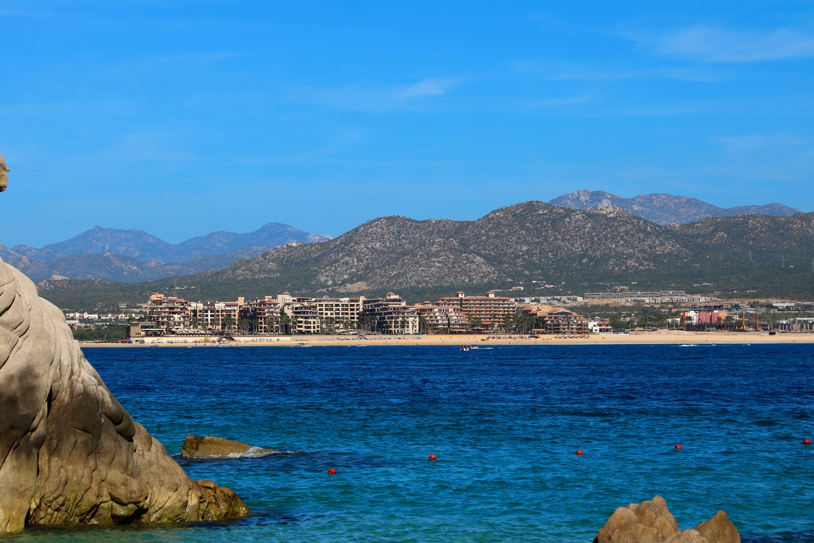 A wide view of Medano Beach in Cabo San Lucas, showing the deep blue water, resorts along the shoreline, and distant mountains.