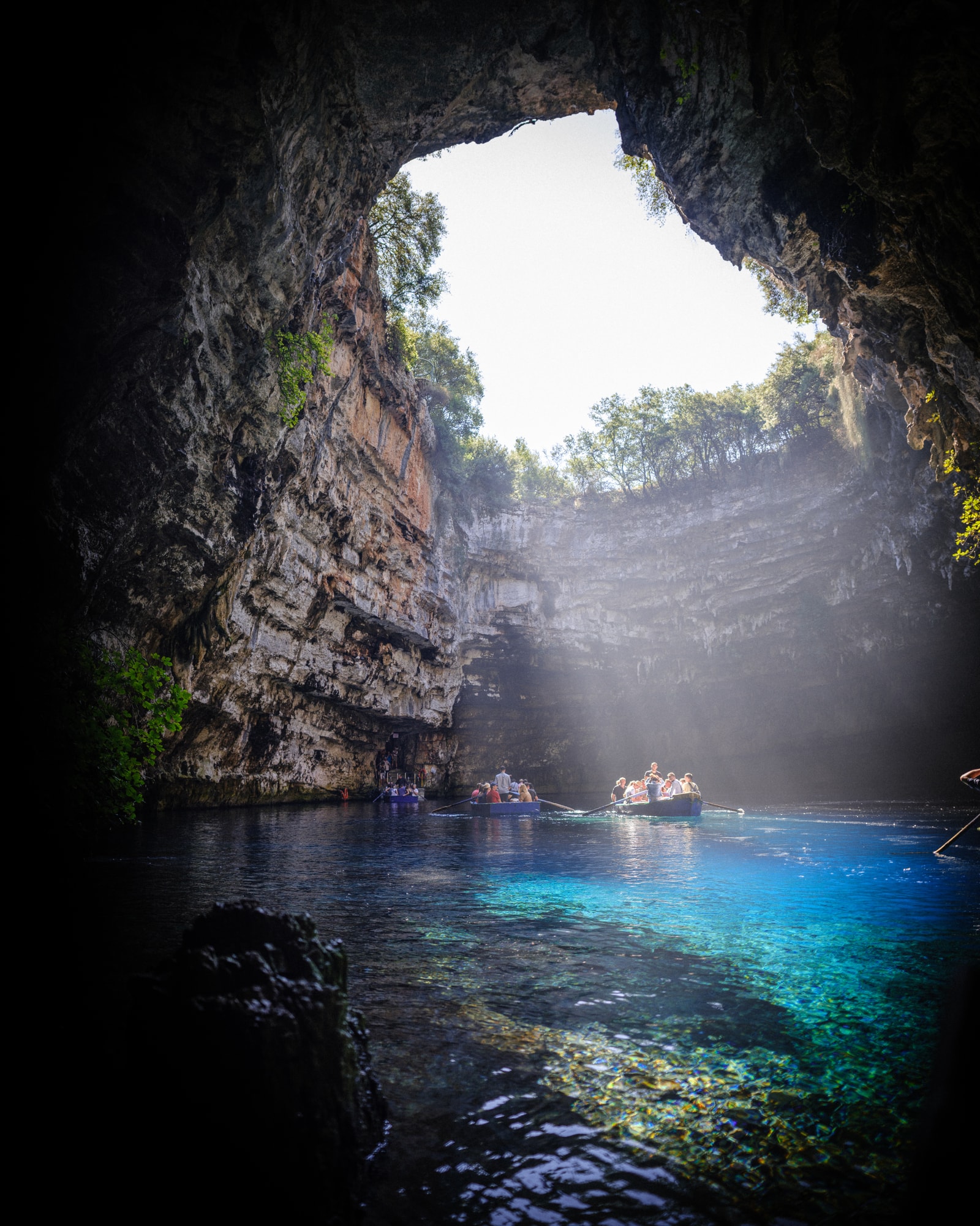 A small rowboat navigates the stunningly clear turquoise water of Melissani Cave Lake as sunlight beams down from the open roof of the cavern.