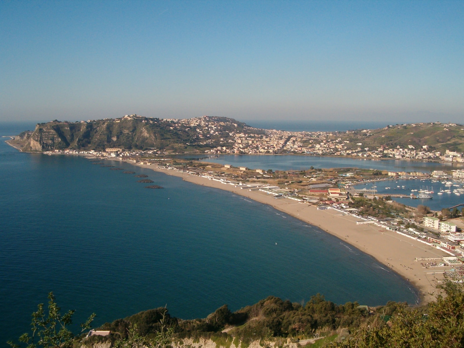 A view of Miliscola beach showing the long strip of dark volcanic sand, the sea, and the towering volcanic cone of Capo Miseno in the background.