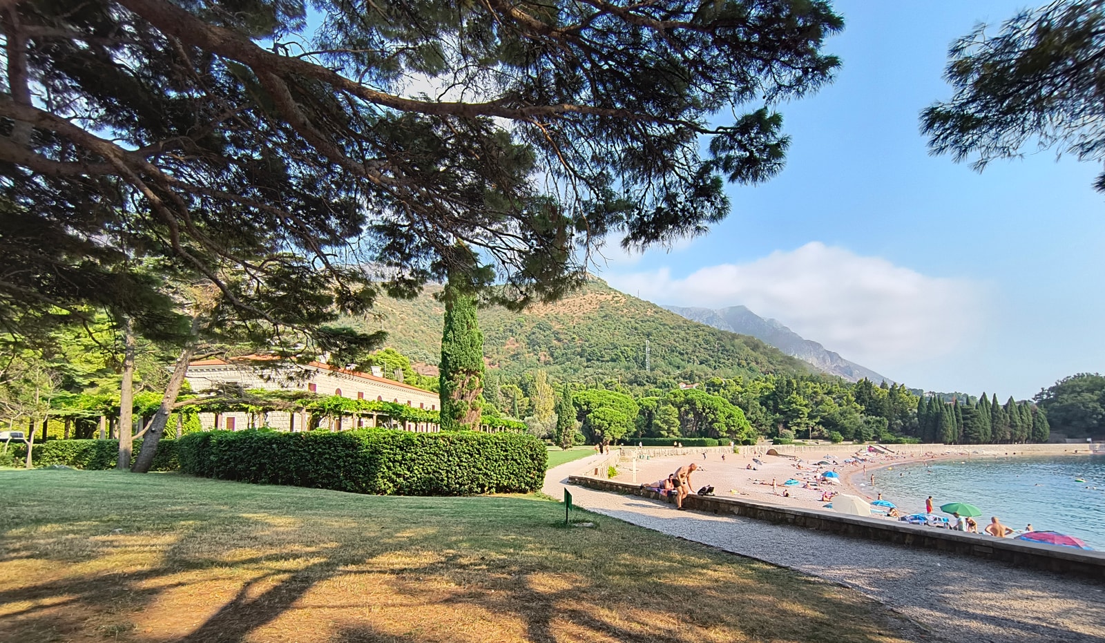A beautiful crescent-shaped beach with calm turquoise water is framed by lush green trees, with a historic stone villa visible in the background.