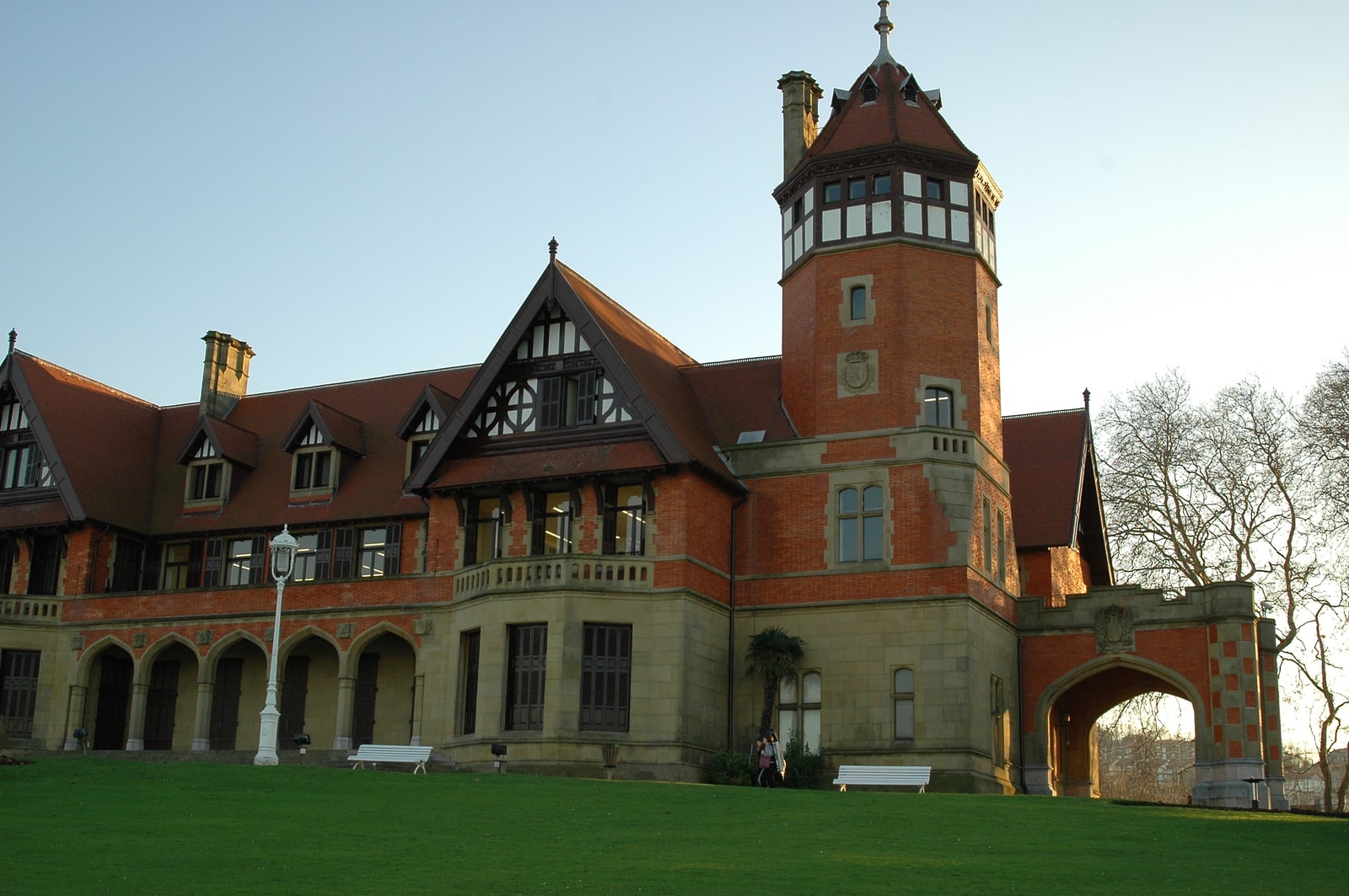 The facade of Miramar Palace, showing its English Tudor style with red brickwork, sandstone trim, and an octagonal turret, set against a green lawn.