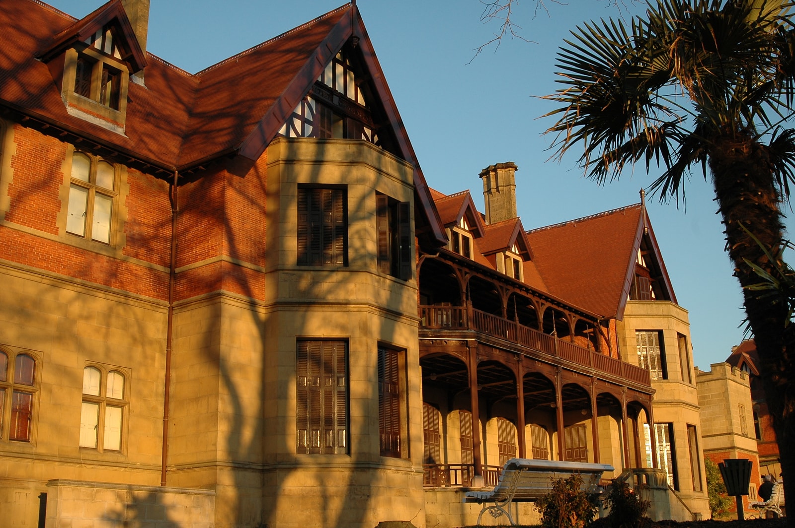 A close-up of Miramar Palace's architecture, showing ornate woodwork, balconies, and brickwork in warm, golden-hour light with palm tree shadows.