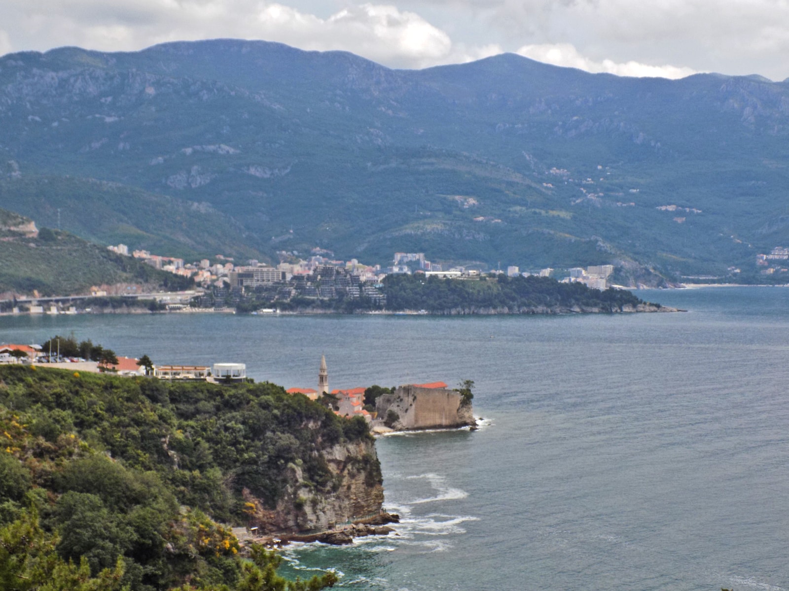 A high-angle view shows a beach cove below a vegetation-covered cliff, with mountains in the distance.