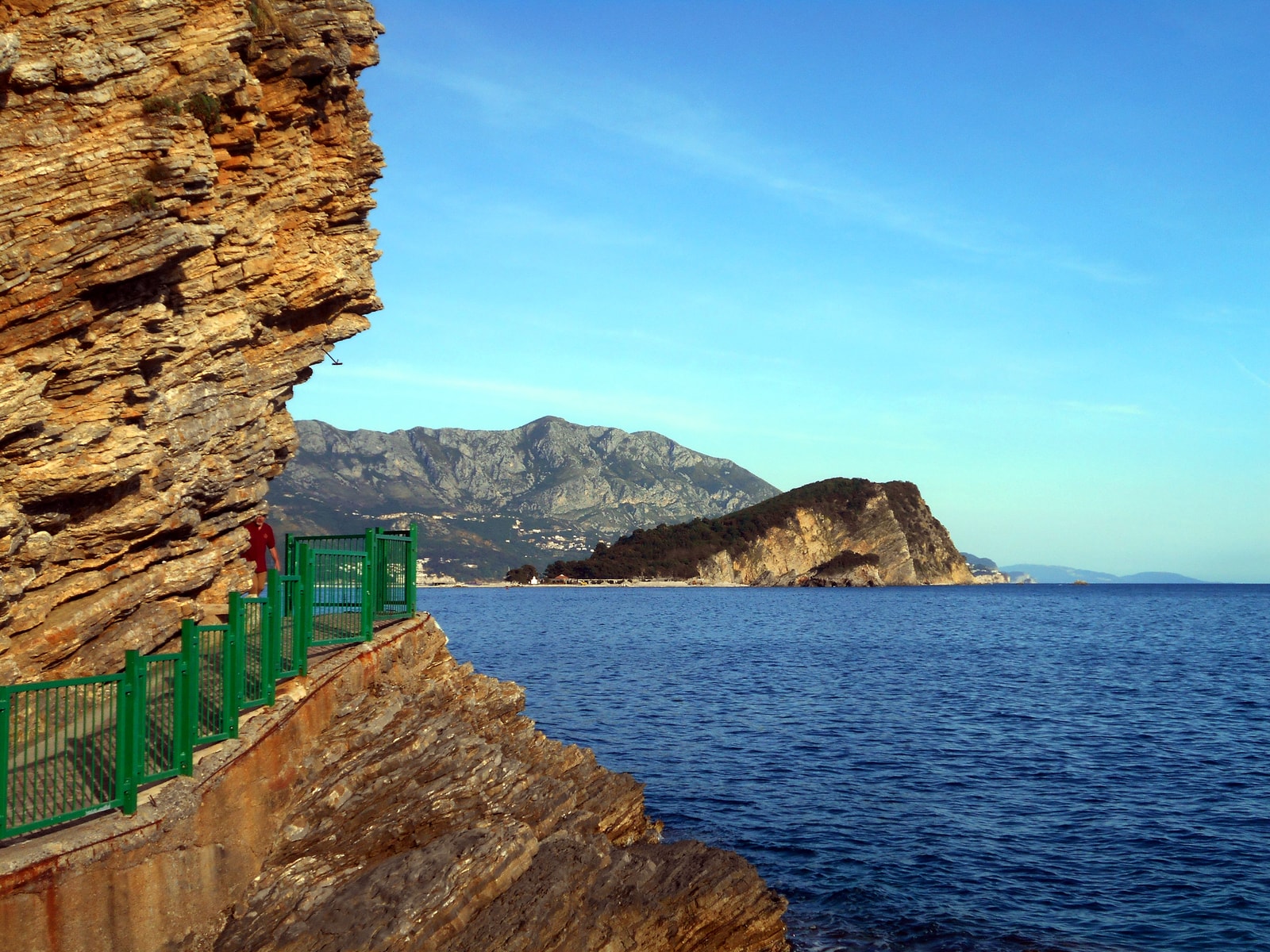 A paved pedestrian path is carved into the side of a sheer, weathered limestone cliff, with metal railings on the seaward side.