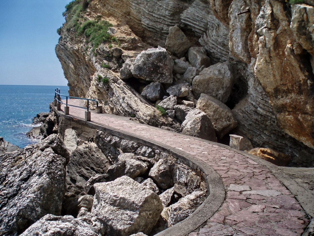 A narrow paved path clings to a rugged limestone cliffside next to the sea.