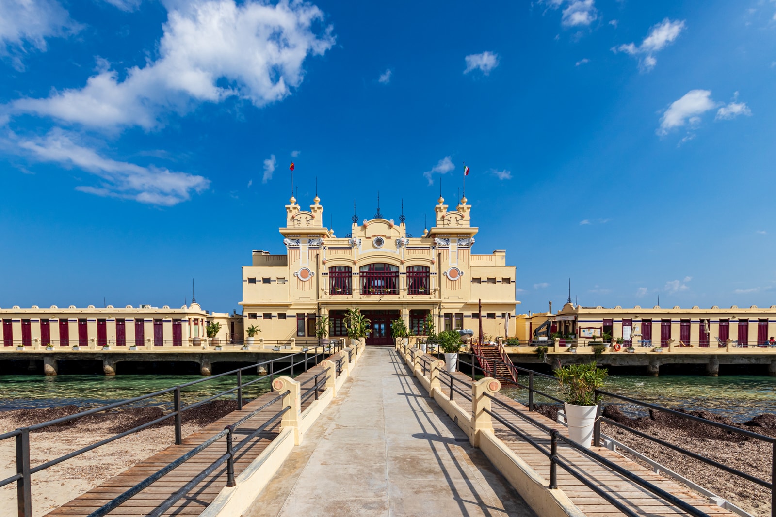 A direct, symmetrical front view of the Antico Stabilimento Balneare, an Art Nouveau structure built on stilts over the turquoise sea under a clear blue sky.