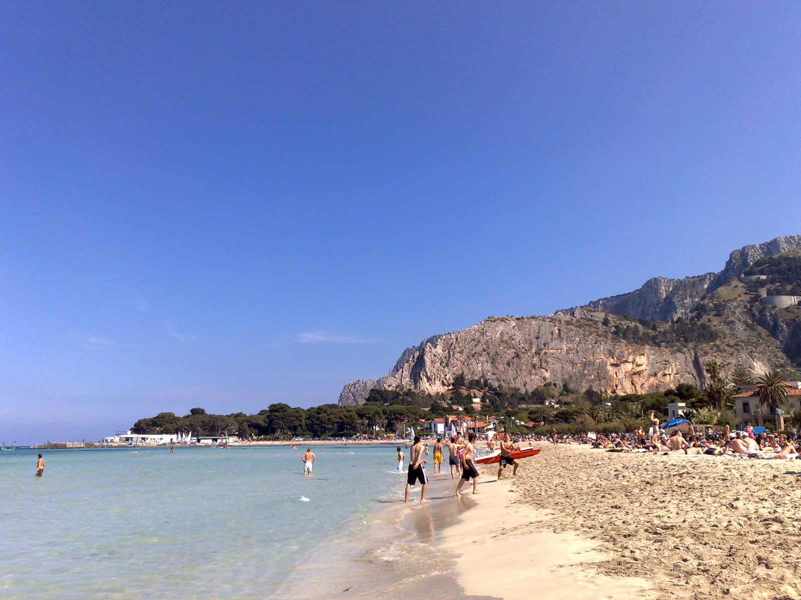 A lively beach scene at Mondello, with numerous sunbathers and swimmers enjoying the clear water under the shadow of a large limestone cliff.