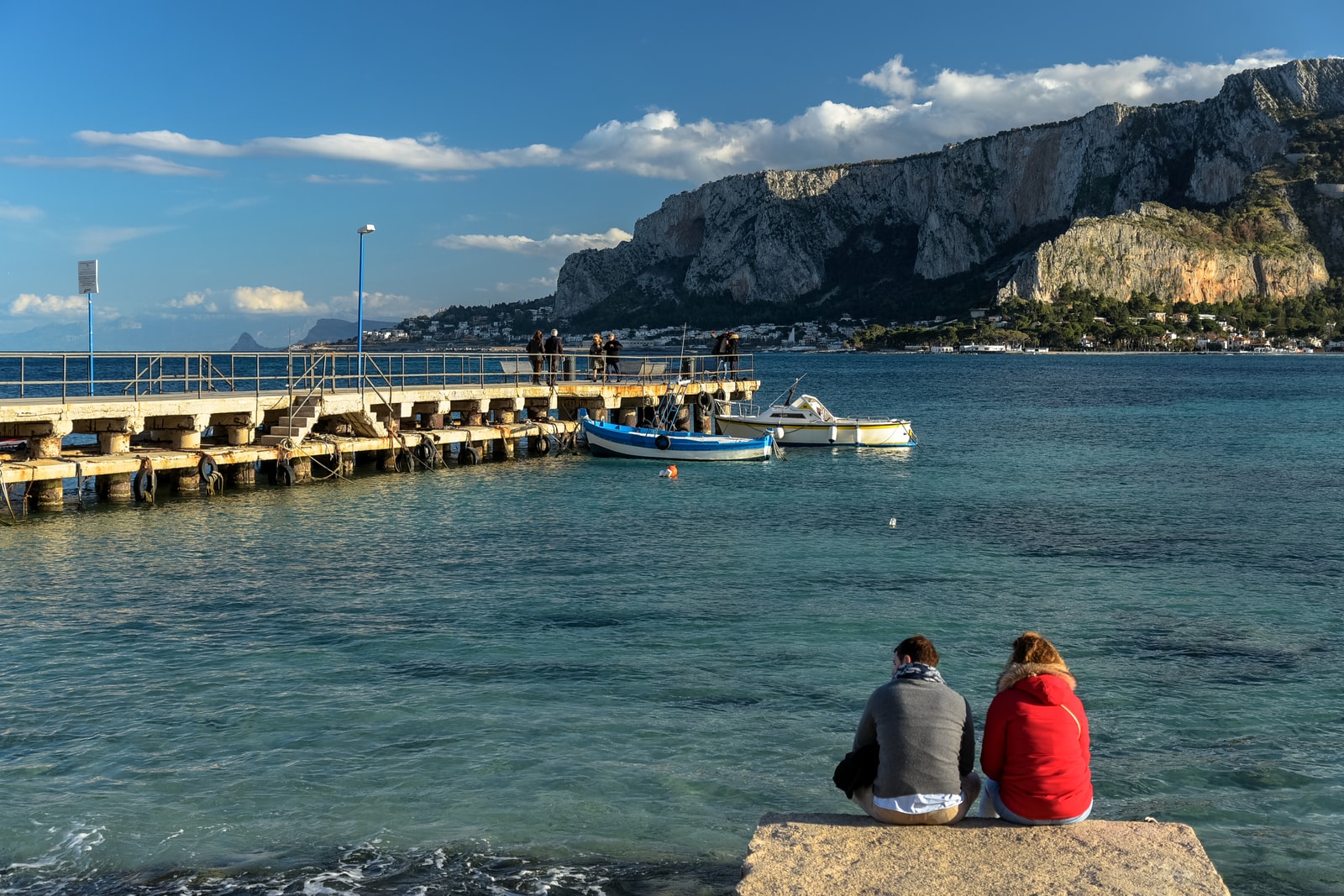 An establishing shot of Mondello, showing the sweep of the bay with sunbathers on a ledge overlooking the beach and mountains.